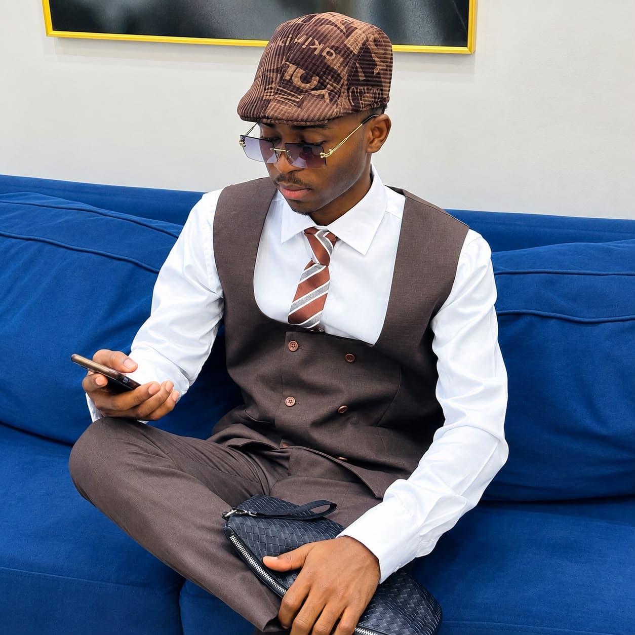 Valdes Banhatio Fosso, assis sur un canapé bleu, en costume marron et casquette, consultant son téléphone